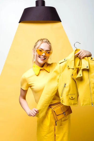 Happy blonde girl in sunglasses holding hanger with jacket on white and yellow — Stock Photo