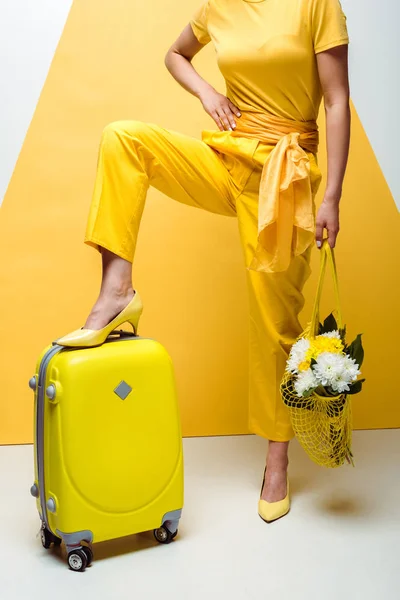 Abgeschnittene Ansicht einer Frau, die mit der Hand an der Hüfte in der Nähe von Gepäck posiert und eine Mehrwegtasche mit Blumen in weiß und gelb hält — Stockfoto