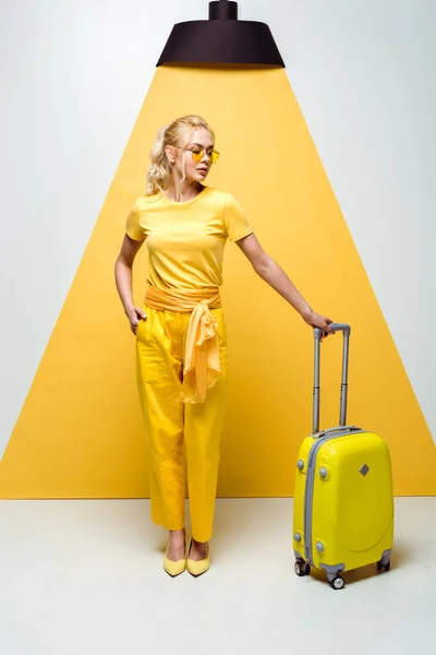 Schöne Frau mit Sonnenbrille posiert mit der Hand in der Tasche und blickt auf Gepäck auf weiß und gelb — Stockfoto