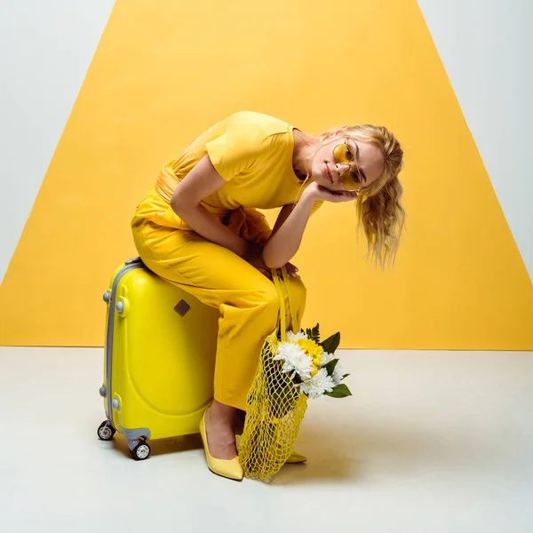 Pretty blonde girl sitting on luggage and holding string bag with flowers on white and yellow — Stock Photo