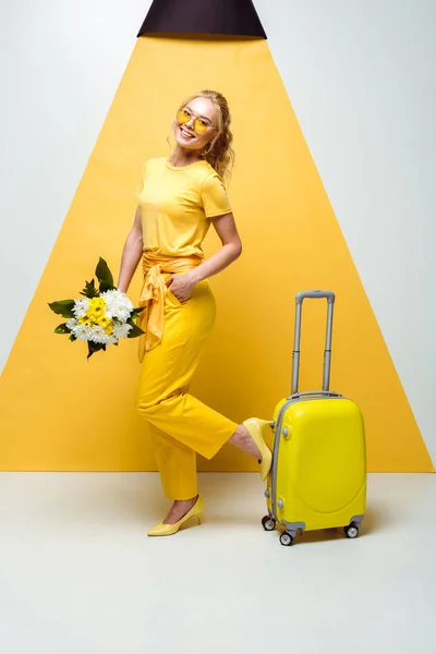 Glückliches blondes Mädchen posiert mit der Hand in der Tasche in der Nähe von Gepäck und hält Blumen auf weiß und gelb — Stockfoto