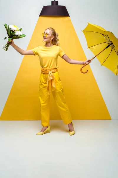 Cheerful blonde girl looking at flowers while holding umbrella on white and yellow — Stock Photo