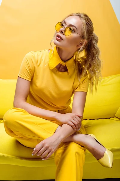 Attraktive blonde Frau mit Sonnenbrille sitzt auf Sofa auf gelb — Stockfoto
