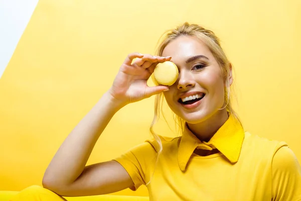 Cheerful blonde girl smiling while covering face with tasty macaron on yellow — Stock Photo
