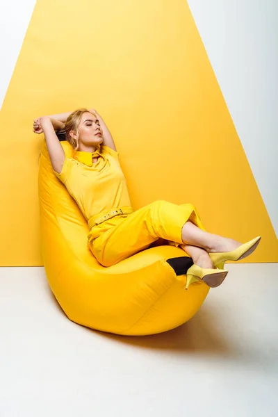 Attractive blonde girl with closed eyes sitting on bean bag chair on white and yellow — Stock Photo