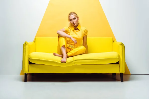 Blonde girl with barefoot sitting on sofa and looking at camera on white and yellow — Stock Photo