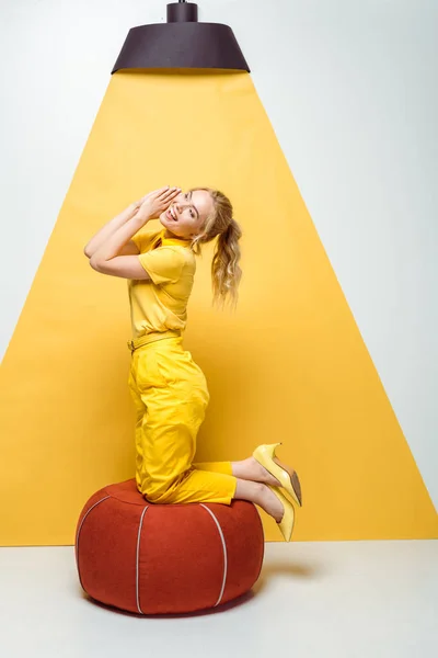 Fröhliches blondes Mädchen lächelt neben rotem Polsterhocker auf weißem und gelbem Grund — Stockfoto