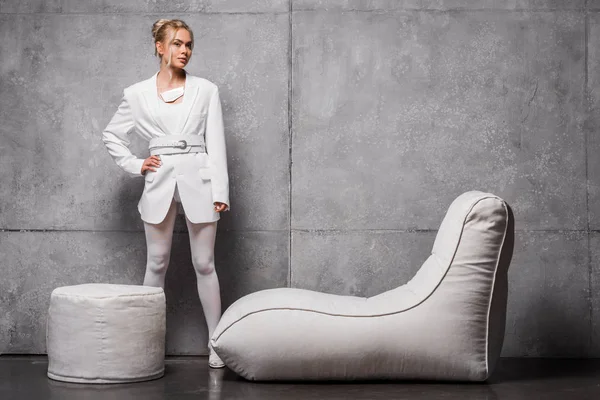 Atractiva mujer rubia posando con la mano en la cadera cerca de la silla bolsa de frijol suave en gris - foto de stock