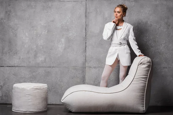 Mujer rubia pensativa posando cerca de la silla bolsa de frijol suave en gris - foto de stock