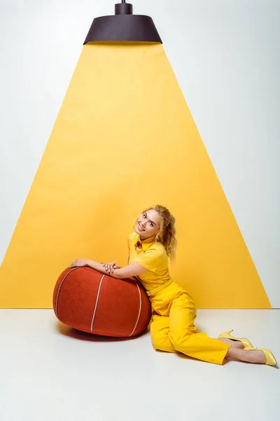 Alegre rubia chica sentado cerca rojo acolchado taburete en blanco y amarillo - foto de stock