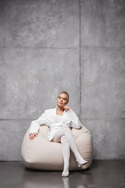 Pensive blonde woman sitting on soft bean bag chair on grey — Stock Photo