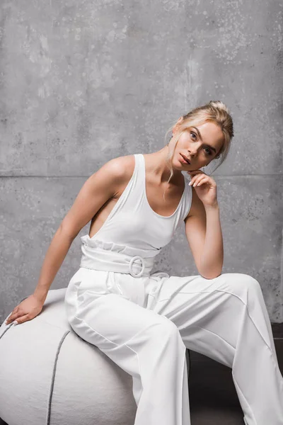 Jolie femme assise sur un tabouret rembourré doux et regardant la caméra sur gris — Photo de stock