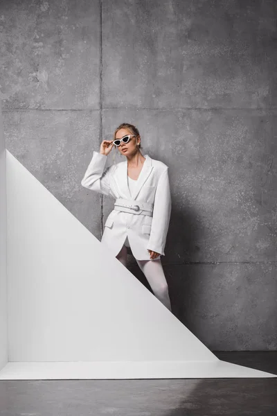 Mulher elegante tocando óculos de sol e posando em branco e cinza — Fotografia de Stock