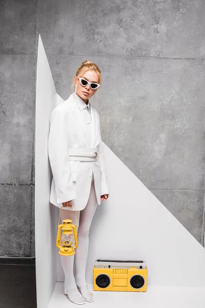 Jolie femme blonde en lunettes de soleil tenant lampe rétro près de boombox jaune sur blanc et gris — Photo de stock
