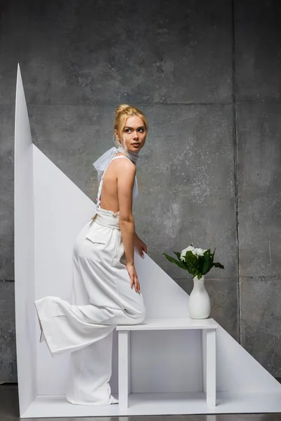Stylish blonde woman posing near vase with flowers on white and grey — Stock Photo