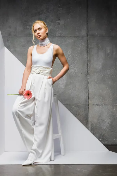 Atractiva mujer posando con la mano en el bolsillo cerca de la silla con flor rosa en blanco y gris - foto de stock