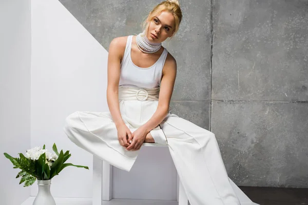 Jolie femme blonde assise près du vase avec des fleurs sur blanc et gris — Photo de stock