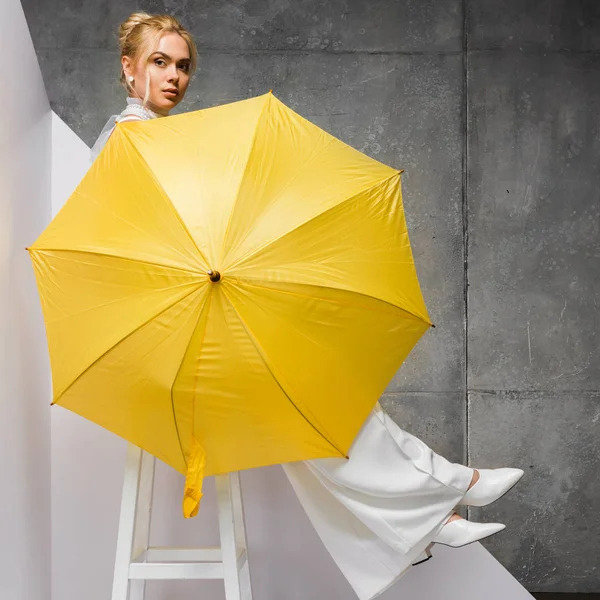 Schönes Mädchen sitzt auf Stuhl und hält gelben Regenschirm auf weiß und grau — Stockfoto