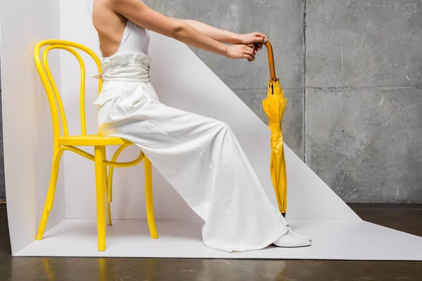 Vue recadrée de la femme assise sur une chaise jaune et tenant un parapluie sur blanc et gris — Photo de stock