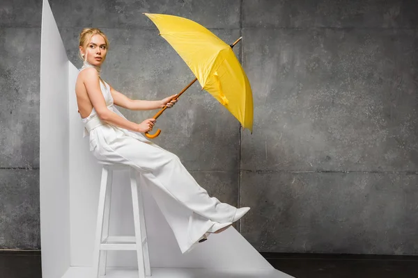 Attraktive blonde Frau sitzt auf Stuhl und hält gelben Regenschirm auf weiß und grau — Stockfoto
