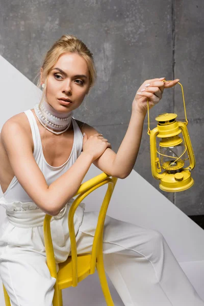 Stylische hübsche Frau sitzt auf gelbem Stuhl und hält Retro-Lampe auf weiß und grau — Stockfoto