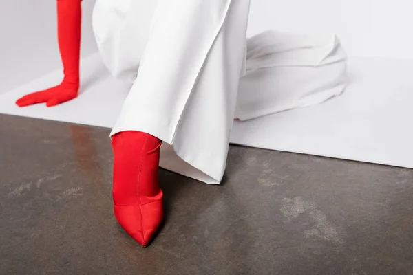 Vista recortada de la mujer joven con estilo en guante rojo y zapato sentado en blanco y gris - foto de stock