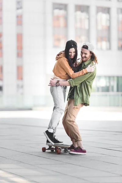 Mulher alegre abraçando com o homem, montando no skate na cidade — Fotografia de Stock
