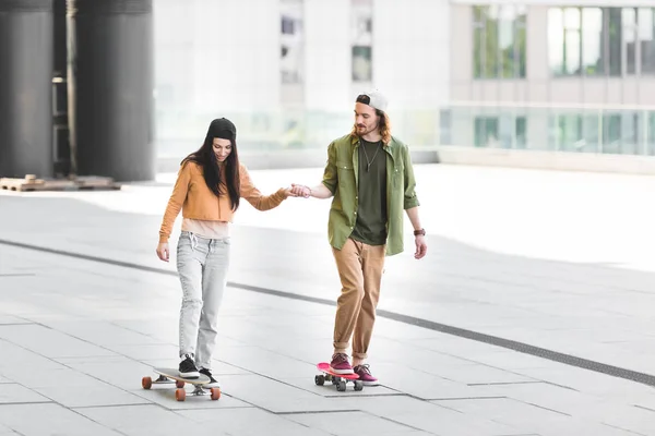 Brunette woman holding hands with man, riding on skateboard in city — Stock Photo