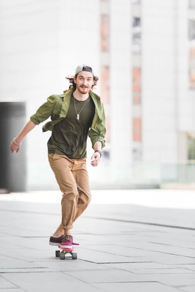Щасливий чоловік дивиться на камеру, їде на скейтборді в місті — стокове фото