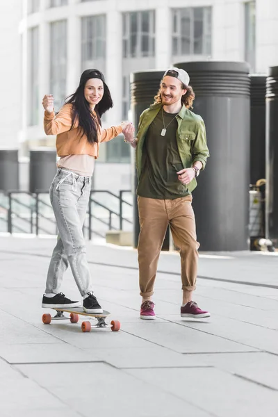 Happy woman holding hands with handsome man, riding on skateboard in city — Stock Photo