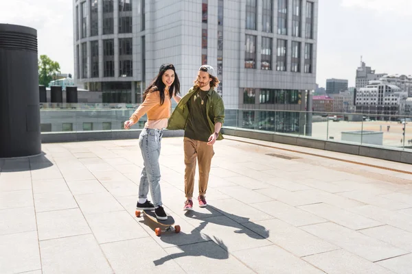 Happy woman holding hands with man, riding on skateboard on roof — Stock Photo