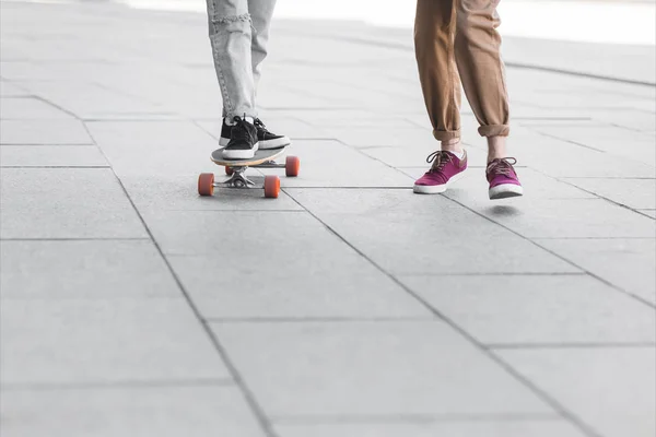 Cropped view of woman in jeans pants riding on skateboard near man — Stock Photo