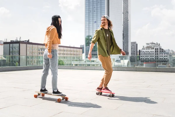Homme en tenue décontractée regardant femme brune, chevauchant sur skateboard sur le toit — Photo de stock