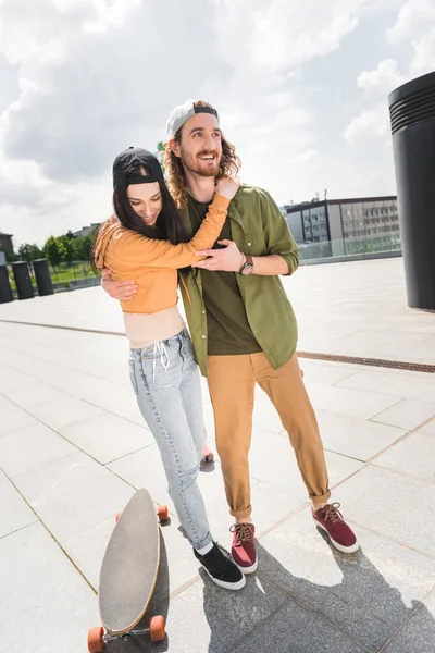 Happy woman hugging handsome man, looking away, standing near skateboard — Stock Photo