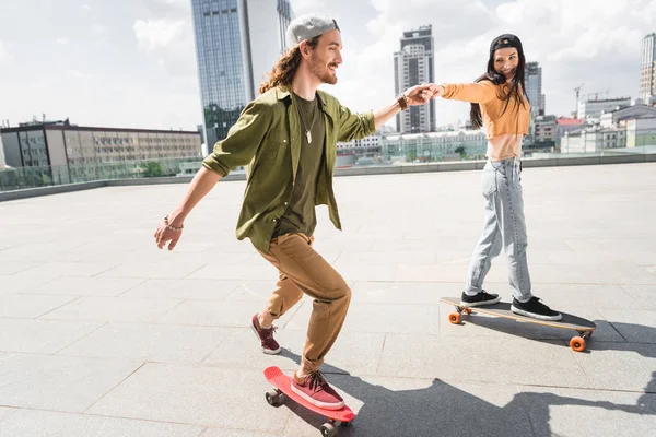 Selektiver Fokus eines glücklichen Mannes, der Händchen hält mit einer schönen Frau, die auf dem Skateboard fährt — Stockfoto