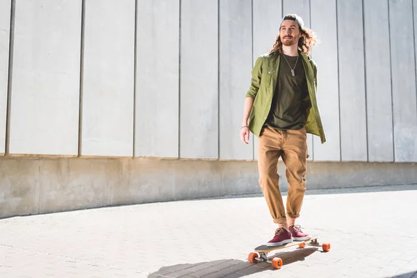 Baixo ângulo de visão de homem bonito olhando para longe, montando no skate perto de parede de concreto — Fotografia de Stock