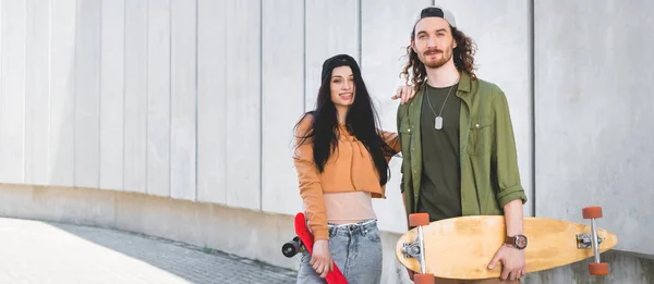 Panoramic view of happy woman in casual wear putting hand on man, standing near concrete wall with skateboards — Stock Photo