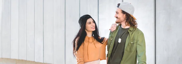 Vista panorámica de la mujer feliz en ropa casual poniendo la mano en el hombre, de pie cerca de la pared de hormigón - foto de stock