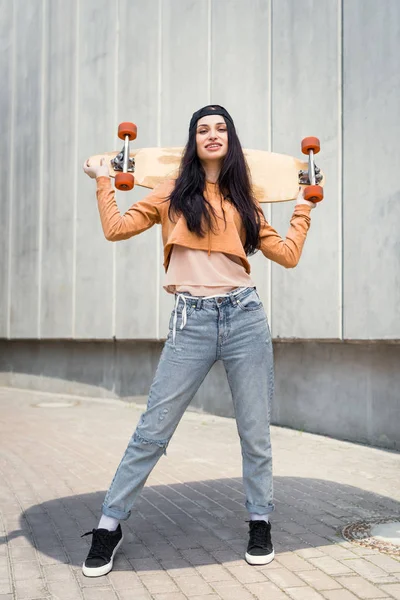 Весела жінка в повсякденному одязі стоїть біля концентрованої стіни, тримаючи скейтборд позаду — Stock Photo