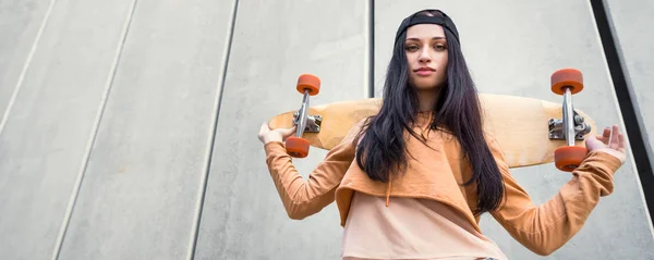 Tiefansicht einer Frau in Freizeitkleidung, die mit Skateboard in der Hand in der Nähe der Wand steht und in die Kamera blickt — Stockfoto