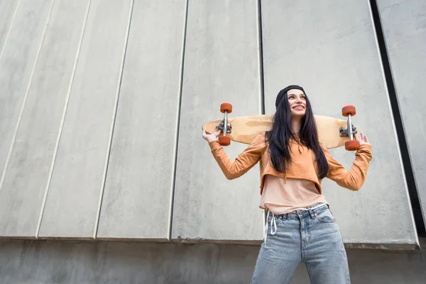 Low angle view of beautiful woman standing near concentrate wall, holding skateboard behind back — Stock Photo