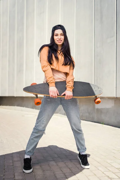 Молода жінка стоїть біля концентрованої стіни, тримаючи скейтборд в руці — Stock Photo