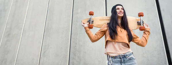 Low angle view of woman in casual wear standing near concentrate wall with skateboard in hands, looking away — Stock Photo
