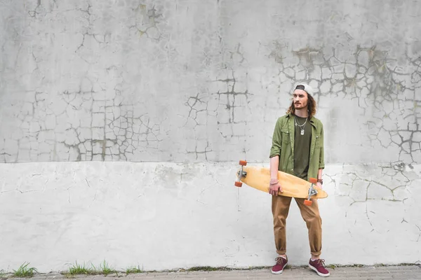 Homem bonito no desgaste casual de pé perto da parede com skate nas mãos — Fotografia de Stock
