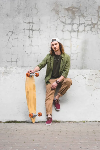 Selbstbewusster Mann in Freizeitkleidung blickt in die Kamera, steht mit Skateboard in der Hand an der Wand — Stockfoto