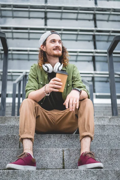 Vista de ángulo bajo de hombre feliz con taza de papel en la mano mirando hacia otro lado, sentado en las escaleras - foto de stock