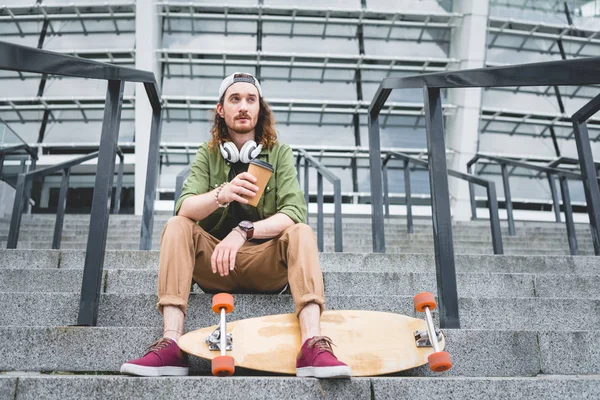 Low angle view of calm man with paper cup in hand looking away, sitting on stairs near skateboard — Stock Photo