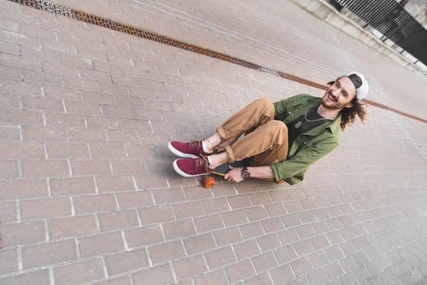 Vista ad alto angolo di uomo allegro seduto e cavalcando sullo skateboard — Foto stock