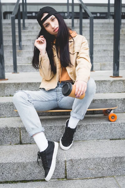 Schöne Frau sitzt auf Skateboard mit Pappbecher in der Hand und blickt in die Kamera — Stockfoto