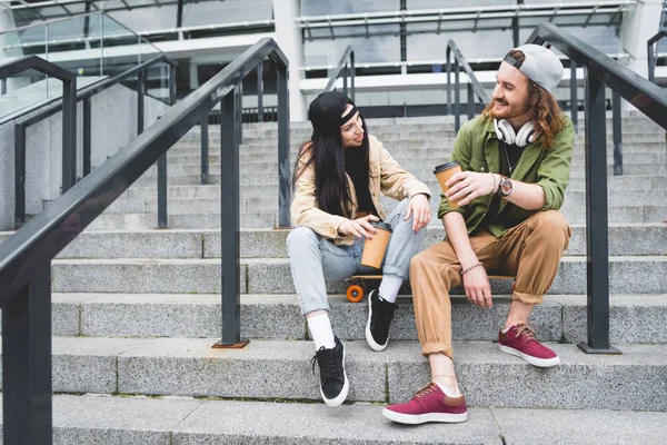 Низкий угол зрения женщины и мужчины, сидящих на скейтборде с бумажной чашкой в руках, глядя друг на друга — стоковое фото